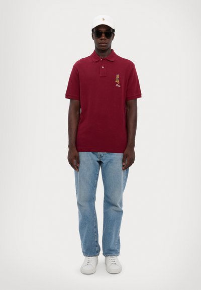 Bordový polo tričko s krátkymi rukávmi a malým multicolor logom. Nosí sa s svetlomodrými džínsami a bielymi teniskami. Biela čiapka a slnečné okuliare.