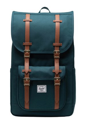 Teal canvas backpack with brown leather straps and gold buckles. Features a front pocket and a branded label at the bottom.