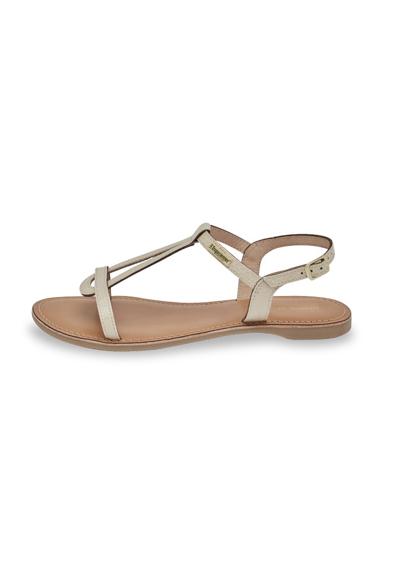 Sandalias planas de cuero beige claro, con un diseño minimalista que presenta correas cruzadas y una correa ajustable en el tobillo. Textura suave, punta descubierta.