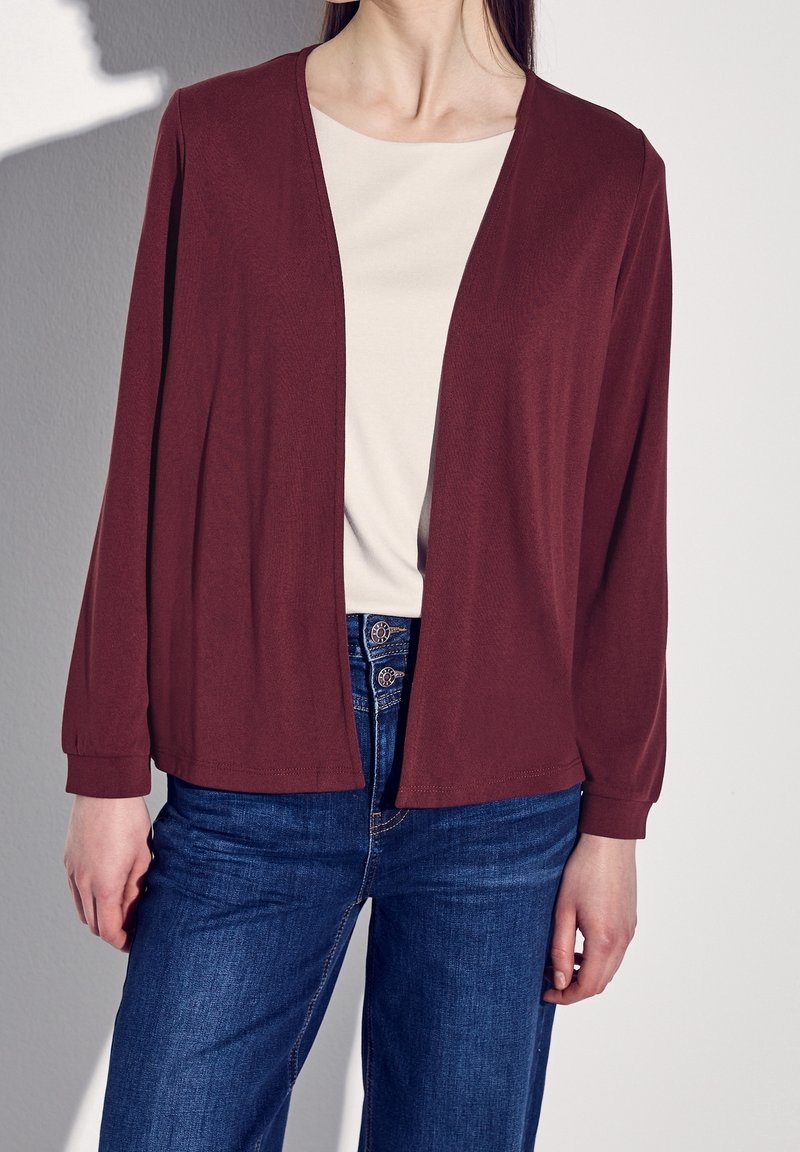 Femme portant un cardigan bordeaux ouvert sur un haut blanc, associé à un jean bleu, debout devant un mur clair uni.
