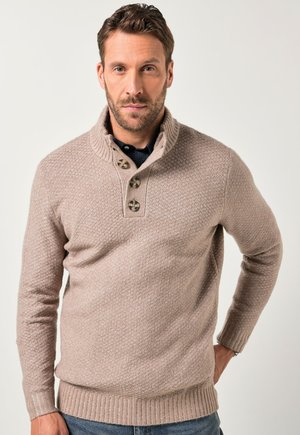 Uomo con capelli corti castani e barba, che indossa un maglione beige a trama con bottoni e jeans blu, in piedi contro uno sfondo semplice.