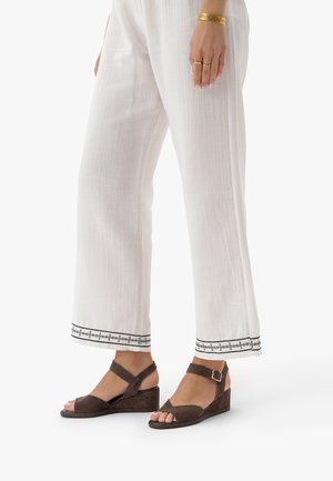 Osoba ubrana w białe spodnie z szerokimi nogawkami z czarną haftowaną lamówką oraz brązowe sandały na koturnie z paskami wokół kostki i złotą klamrą.