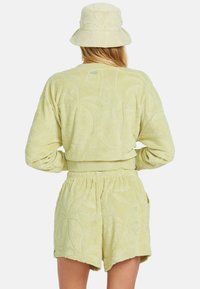 Gelb-grünes Frottee-Outfit mit einem taillierten Langarmoberteil und passenden Shorts mit Palmenblattstruktur, ergänzt durch einen Bucket Hat.