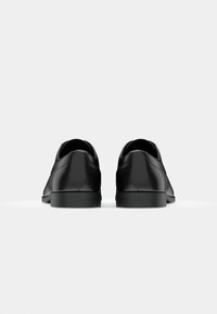 Zapatos de cuero negro con un diseño elegante, punta redondeada y una textura suave. Incluyen una suela de goma negra baja y detalles de costura mínimos.