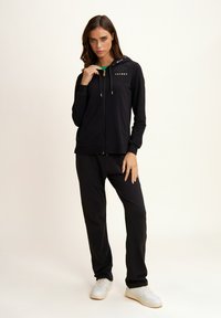 Sudadera negra con cremallera y detalles en dorado, acompañada de pantalones negros a juego. Ambos están hechos de un material suave y elástico.