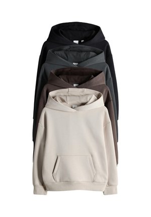 Osmijehno dijete s kosom do ramena, odjeveno u bež hoodicu, plave široke traperice i smeđe natikače stoji uz bijeli zid.