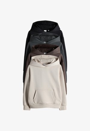 Lachend kind met schouderlang haar, een beige hoodie, blauwe wijde spijkerbroek en bruine instapschoenen, staand tegen een witte muur.