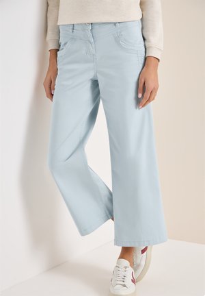 Femme portant un pantalon large bleu clair, un haut à manches longues beige et des baskets blanches avec des accents rouges, debout contre un mur blanc.