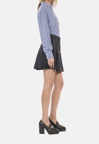 Blau-weiße gestreifte Bluse mit langen Ärmeln, kombiniert mit einem schwarzen plissierten Rock mit goldenen Knöpfen und schwarzen Absatzslippern.