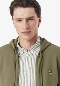 Felpa verde oliva con zip, con una tasca a marsupio, indossata sopra una camicia button-up bianca e verde a righe, caratterizzata da un piccolo logo Lacoste.