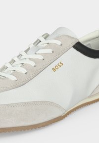 Λευκό και μπεζ δερμάτινο sneaker με λευκά κορδόνια, μπεζ σουέντ λεπτομέρειες, μαύρη διακόσμηση στη φτέρνα και χρυσό λογότυπο "BOSS" στο πλάι.