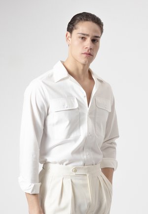 Joven con cabello oscuro peinado hacia atrás que lleva una camisa blanca abotonada y pantalones plisados crema, de pie contra un fondo claro y liso.