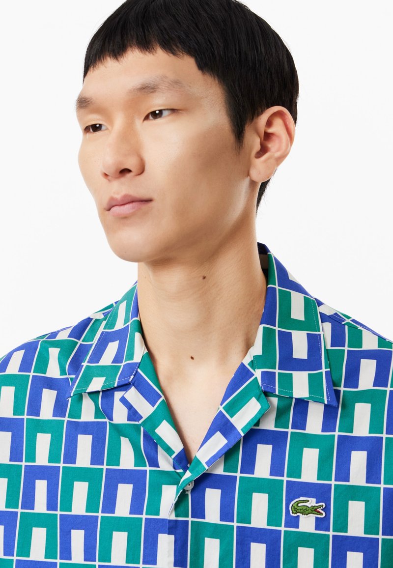 Chemise à manches courtes avec un motif géométrique vert et bleu, ornée de carrés blancs et d'un logo brodé sur la poitrine.