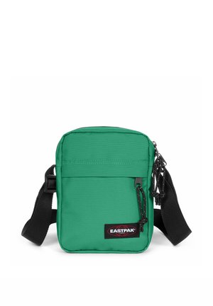 Pequeña bolsa de hombro Eastpak verde con correa negra ajustable, bolsillo delantero con cremallera y parche con logo sobre fondo blanco.