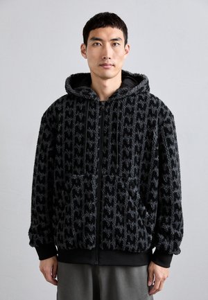 Zwarte fuzzy capuchonjack met een geprofileerd patroon, voorzakken en een ritssluiting. De manchetten en taille zijn van effen zwart.