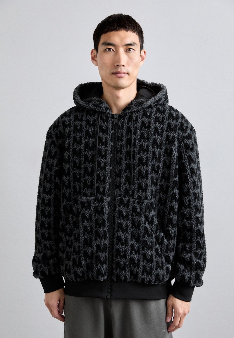 Black fuzzy hooded jacket with a textured pattern, front pockets, and a zipper closure. Cuffs and waist trim are solid black.