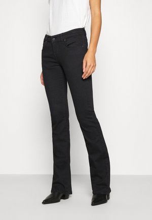 Kobieta w czarnych dżinsach bootcut, białym bezrękawnym topie i czarnych botkach z ostrym noskiem, stojąca na jednolitym tle.