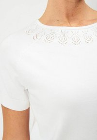 Valkoinen lyhythihainen neulepaita, jossa on teksturoitu kaulus ja pieniä koristekuvioita, pyöreä kaula-aukko sekä resorit hihansuissa. Sujuva kankaan rakenne.