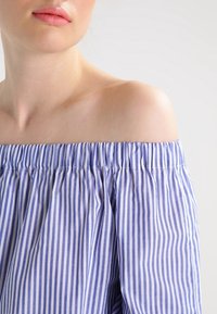Blusa fuera del hombro en azul claro y blanco con rayas verticales, que presenta una textura fruncida en el escote y una tela fluida.
