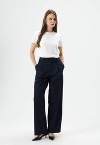 T-shirt blanc à manches courtes associée à un pantalon large bleu marine, avec des plis à l'avant et des poches latérales. Des talons noirs à bout pointu complètent la tenue.