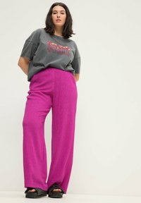 Grijze oversized t-shirt met rode en gele tekst, gecombineerd met felroze wijde broek met een textuurafwerking en zwarte platformsandalen.