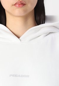 Hvit hettegenser med frontlomme og snor i hetten. Merkenavnet "PEGADOR" er trykket i lys grå på brystområdet.