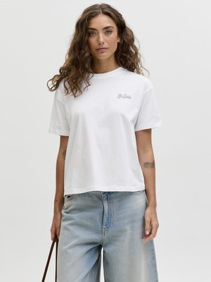 Kvinde med bølget hår iført en hvid "La Siesta" T-shirt og lyseblå jeans, der holder en brun rem i venstre hånd og står foran en ensfarvet baggrund.