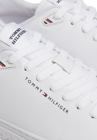 Sneakers in materiale sintetico bianco con lacci bianchi piatti. Presentano un'etichetta con logo intrecciato e il nome del marchio stampato in nero con accenti rossi e blu.