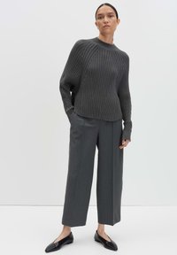 Donna che indossa un maglione a coste grigio scuro, pantaloni a gamba larga abbinati e scarpe nere pianeggianti, in piedi contro uno sfondo chiaro e semplice.