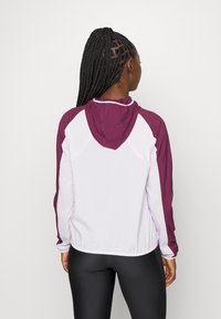 Veste légère zippée avec un corps blanc et des manches violet foncé, dotée d'une capuche et d'un ourlet élastique. Texture lisse, design sportif.