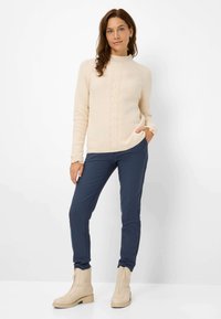 Maglione beige a maglia con collo alto e motivo a treccia, abbinato a pantaloni fitted blu navy e stivaletti crema con zip laterali.