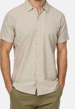 Man wearing a beige short-sleeve button-up shirt with collar and olive green pants, hands in pockets, against white background.