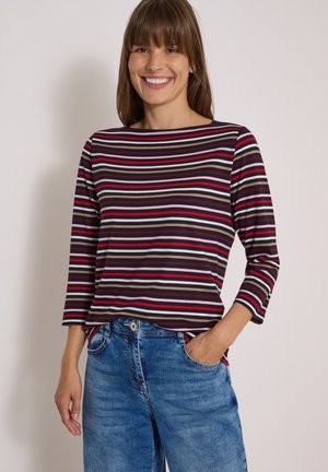 Femme souriante portant un haut rayé à manches longues avec des lignes rouges, noires, blanches et beiges et un jean bleu, une main dans la poche.