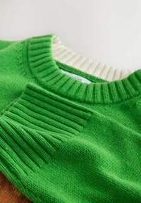 Zaļa adīta džempera augšdaļa ar ribu izgriezumu un teksturētām akcentiem, ar kontrastējošu beidzējo krēslas detaļu.