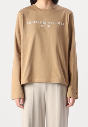 Person iført en brun langærmet skjorte med teksten "Tommy Hilfiger Est. 1985" og beige bukser, stående foran en ensfarvet baggrund.