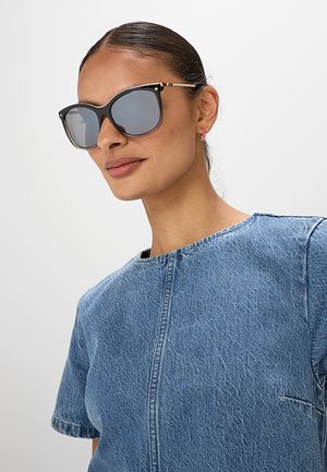 Donna con occhiali da sole con montatura nera, orecchini a cerchio rosa e top corto in denim blu su sfondo chiaro uniforme.