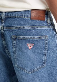Jeans de denim en un color azul desgastado, con una etiqueta de cuero marrón y un parche de logo triangular blanco en el bolsillo trasero.