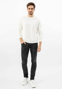 Weißes Langarmhemd, schwarze Jeans und weiße Sneaker. Schmale Passform, weicher Stoff und sauberes Design. Mann steht mit Händen in den Taschen vor neutralem Hintergrund.