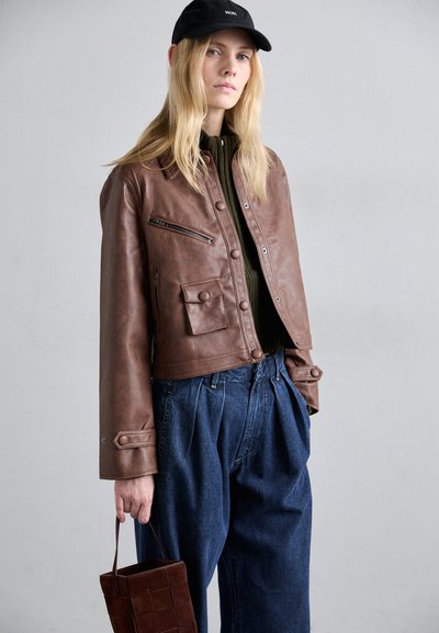 Brun læder cropped jakke med en lynlåslomme, parret med løstsiddende blå denim bukser og en lille vævet taske. Sort kasket på hovedet.