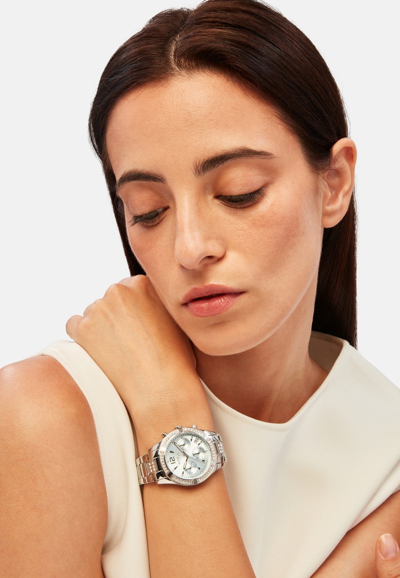 Montre chronographe en argent avec un cadran rond, trois compteurs, affichage de la date, et un bracelet en métal texturé, portée sur un poignet avec une peau lisse.