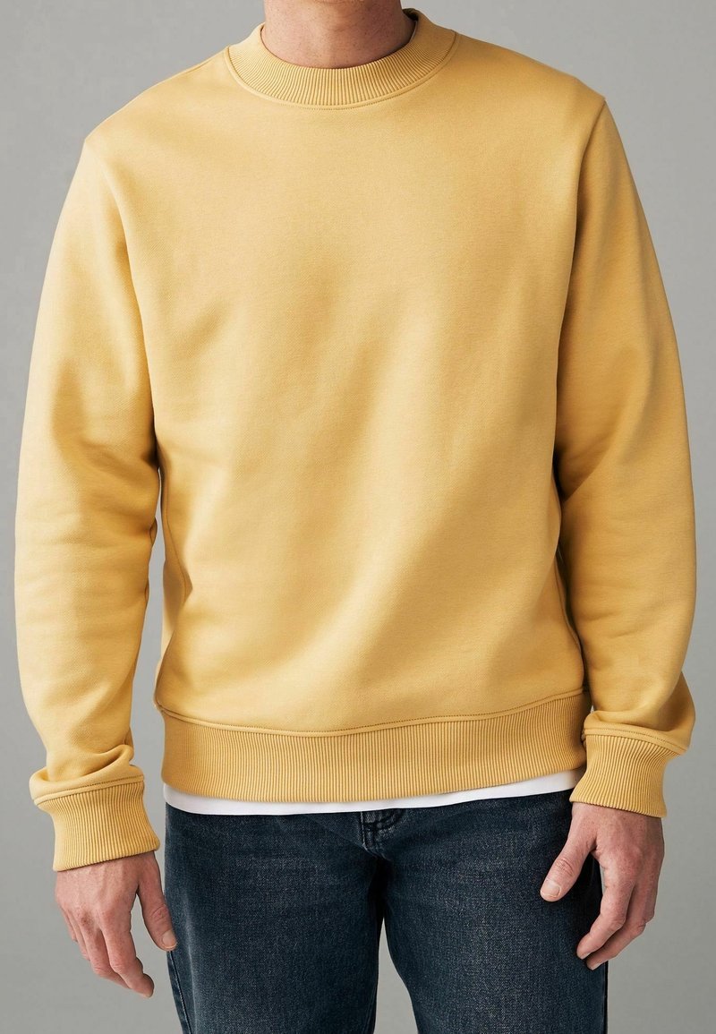 Homme portant un sweat-shirt jaune uni à col rond par-dessus une chemise blanche et un jean bleu foncé, debout devant un fond gris.