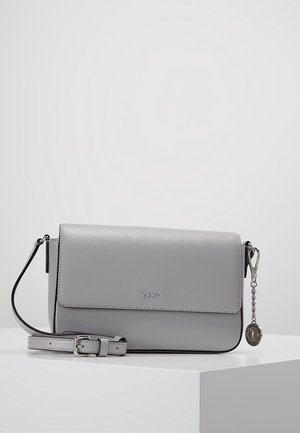 Sac à main DKNY en cuir gris clair avec bandoulière réglable, fermeture à rabat et chaîne argentée attachée avec un pendentif rond, posé sur une surface blanche.