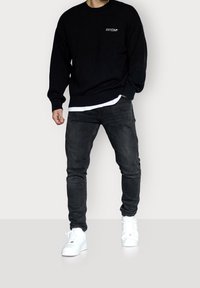 Pull en maille noire avec poignets côtelés, porté sur une chemise blanche, associé à un jean foncé et des baskets blanches. Accent mis sur le confort et un style décontracté.