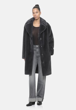 Donna con i capelli raccolti a chignon, che indossa un ampio cappotto in pelliccia sintetica nera, una maglietta nera trasparente, pantaloni grigi arrotolati e stivaletti neri con tacco, in piedi su sfondo bianco.