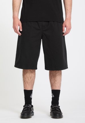 Homme portant un short noir jusqu'aux genoux, des chaussettes noires avec un logo blanc, des mocassins noirs et un t-shirt noir sur fond blanc.