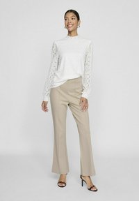 Top blanco de manga larga con patrón texturizado, cuello alto y mangas transparentes, combinado con pantalones de pierna ancha beige y sandalias de tacón negro.