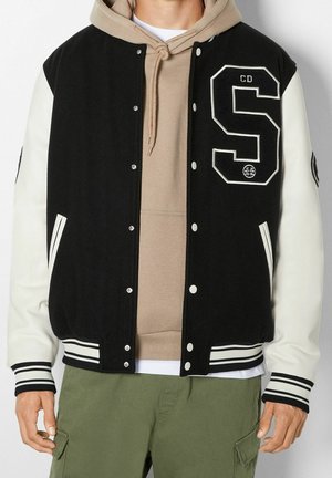 Homme portant une veste letterman noire et blanche avec un grand écusson "S", un sweat à capuche beige et un pantalon cargo vert olive sur un fond uni.
