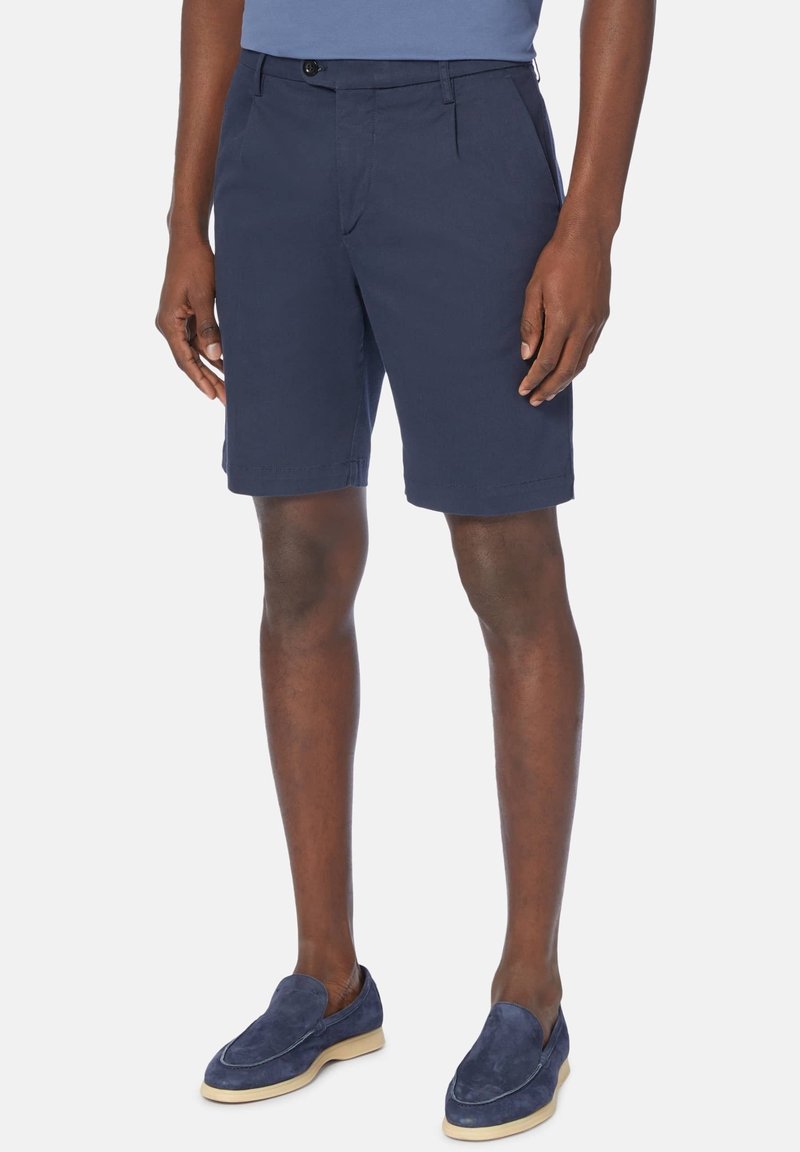 Homme portant un short bleu marine sur mesure et des mocassins en daim assortis, debout devant un fond uni.