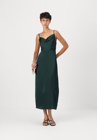 Mörkgrön satinmidiklänning med cowl-urringning, tunna axelremmar och en figurnära midja, matchad med svarta sandaler och en liten metallisk clutch.
