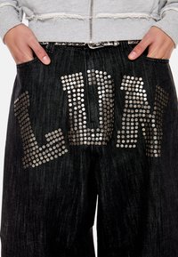 Χέρια στις τσέπες μαύρων τζιν με ασημένια γράμματα "LUCK" με τρουκς, σε συνδυασμό με γκρι φούτερ με φερμουάρ.